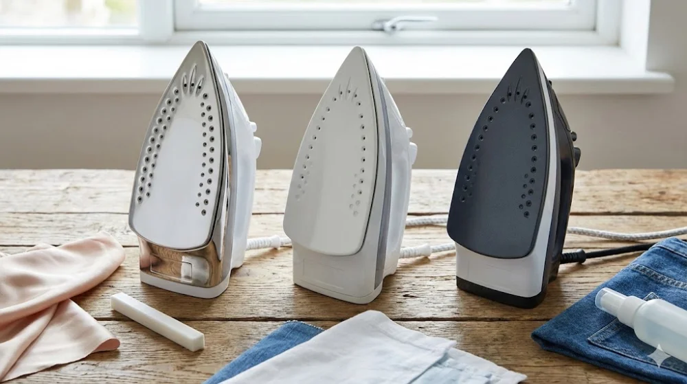 Три праски з підошвами з нержавійки, кераміки та тефлону на дерев'яному столі зі зразками тканин і засобами догляду